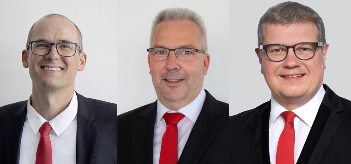 Drei Männer in Anzügen und roten Krawatten lächeln in die Kamera.