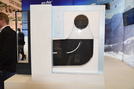 Ein weißes Gerät mit einem runden Fenster steht auf einem Tisch, im Hintergrund sind Menschen und ein Banner mit dem Logo 