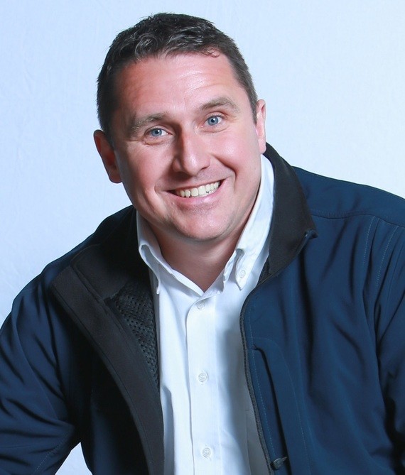 Ein Mann mit einem Lächeln trägt ein weißes Hemd und eine dunkelblaue Jacke.