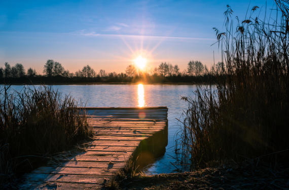 Ein Holzsteg führt zu einem ruhigen See, während die Sonne am Horizont aufgeht und den Himmel in warme Farben taucht.