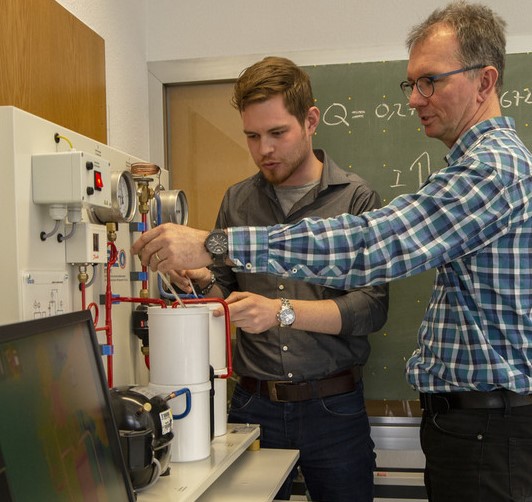 Zwei Personen arbeiten gemeinsam an einem Laborapparat, während eine Tafel mit mathematischen Formeln im Hintergrund zu sehen ist.