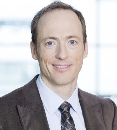 Ein Mann mit kurzen blonden Haaren trägt einen dunklen Anzug, ein weißes Hemd und eine karierte Krawatte.