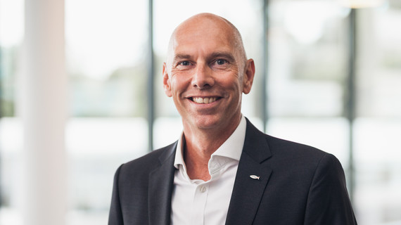 Ein Mann im Anzug lächelt in die Kamera vor einem hellen Hintergrund.