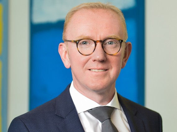 Ein Mann im Anzug und Krawatte lächelt in die Kamera vor einem blauen Hintergrund.