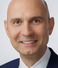 Ein Mann mit kurzem Haar und einem Lächeln trägt einen dunklen Anzug mit weißem Hemd.