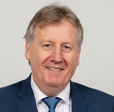 Ein Mann mit grauem Haar und einem Lächeln trägt einen dunkelblauen Anzug mit einer hellblauen Krawatte.