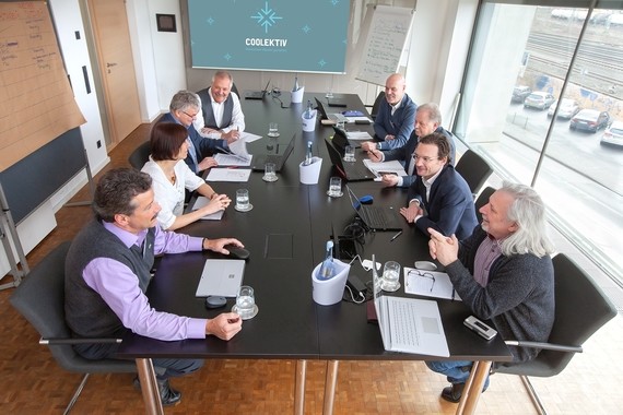 Eine Gruppe von Menschen sitzt um einen Konferenztisch und führt ein Meeting in einem modernen Büro mit großen Fenstern im Hintergrund.