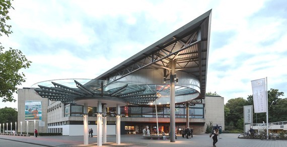 Ein modernes Gebäude mit einer großen, schattierten Dachkonstruktion und einer klaren Fassade, umgeben von Bäumen und Menschen, die sich im Freien aufhalten.