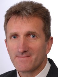 Ein Mann mit kurzen, hellbraunen Haaren und einem ernsten Gesichtsausdruck, der in ein helles Licht blickt.