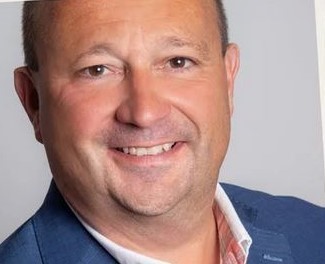 Ein Mann mit kurzen, dunklen Haaren und einem Lächeln trägt einen blauen Anzug und ein weißes Hemd.