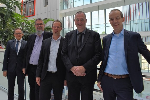 Fünf Männer in Anzügen stehen nebeneinander in einem modernen Bürogebäude.