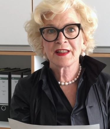 Eine ältere Frau mit Brille und Perlenkette sitzt an einem Schreibtisch und betrachtet Dokumente.