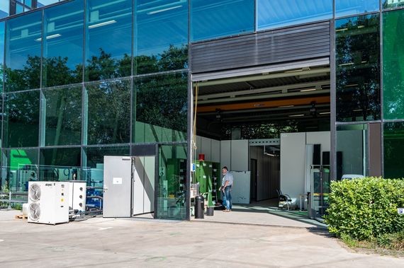 Ein modernes Bürogebäude mit großen Fensterfronten und einer offenen Einfahrt, umgeben von Grünflächen und einigen Personen im Vordergrund.