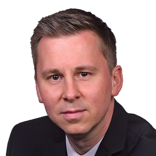 Ein Mann mit kurzem dunklem Haar und Bart trägt ein dunkles Hemd mit Krawatte.