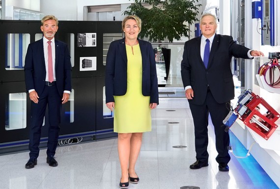Drei Personen stehen nebeneinander in einem modernen Büro, wobei zwei Männer in Anzügen und eine Frau in einem gelben Kleid zu sehen sind.