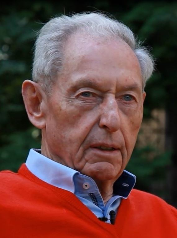 Ein älterer Mann mit grauem Haar trägt einen roten Pullover über einem blauen Hemd.