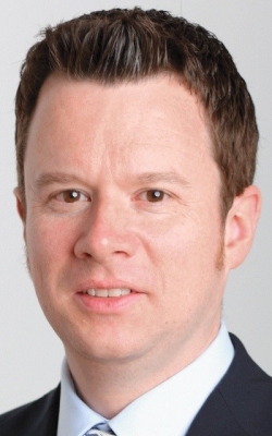 Ein Mann mit kurzen dunklen Haaren und einem Lächeln trägt ein weißes Hemd und eine dunkle Krawatte.