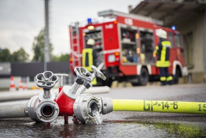 Ein Feuerwehrauto steht im Hintergrund, während im Vordergrund ein Feuerlöschschlauch mit Wasser spritzt.
