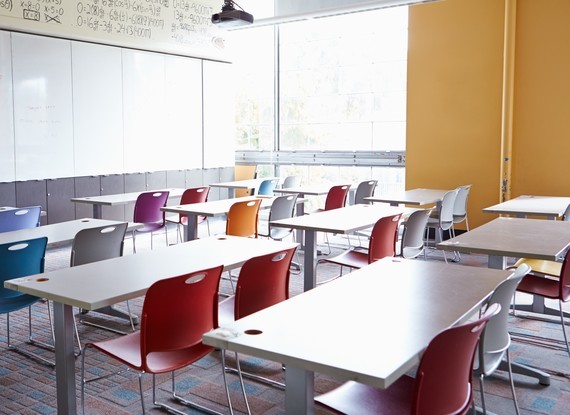 Ein leeres Klassenzimmer mit weißen Tischen, bunten Stühlen und großen Fenstern.