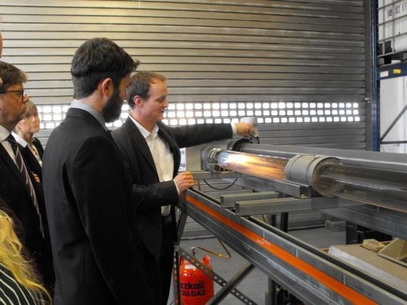 Drei Personen in Anzügen stehen in einer Werkstatt und betrachten einen großen Metallzylinder auf einer Werkbank.