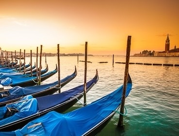 Reihe von Gondeln mit blauen Abdeckungen am Ufer eines Kanals bei Sonnenuntergang.