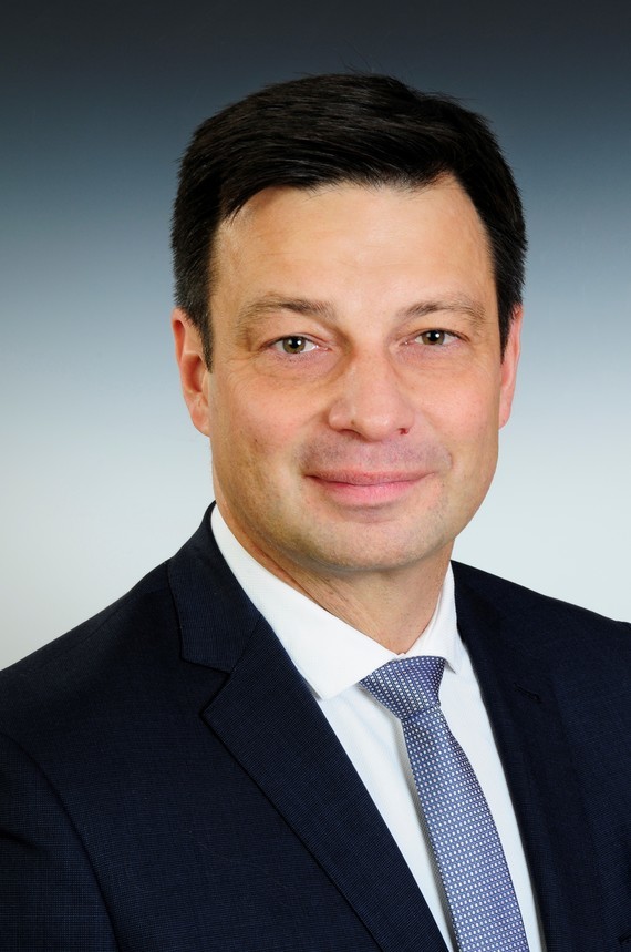 Ein Mann mit dunklem Haar und Bart trägt einen dunklen Anzug, ein weißes Hemd und eine blaue Krawatte.