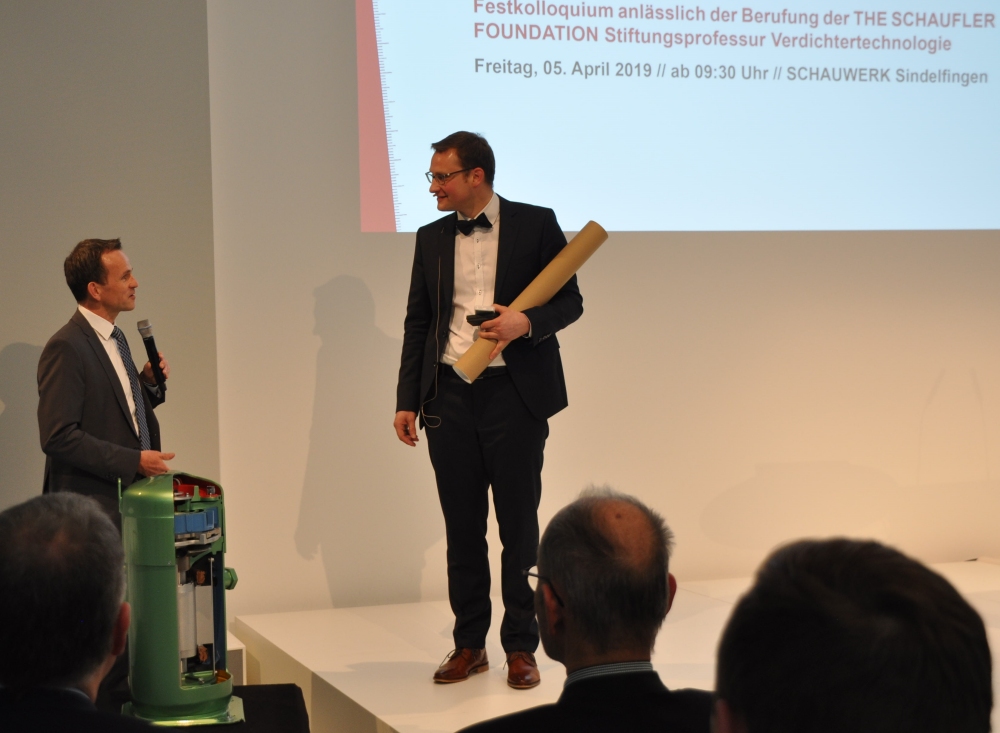 Zwei Männer stehen auf einer Bühne, einer hält einen Hammer, während im Hintergrund ein Präsentationsbildschirm mit Text zu sehen ist.