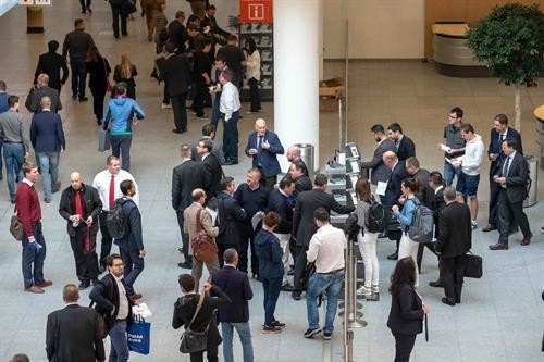 Ein belebter Innenraum mit vielen Menschen, die sich unterhalten und sich bewegen, während im Hintergrund ein großer Pfeiler und Pflanzen zu sehen sind.