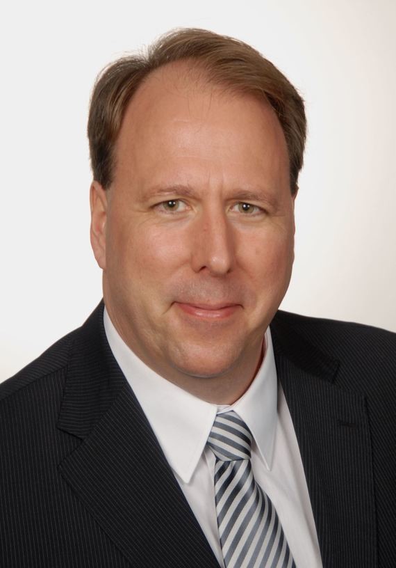 Ein Mann mit kurzem braunem Haar trägt einen dunklen Anzug, ein weißes Hemd und eine gestreifte Krawatte.