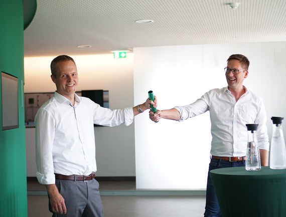 Zwei Männer stehen in einem hellen Raum und schütteln sich die Hände, während einer eine grüne Flasche hält.