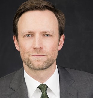 Ein Mann mit kurzen blonden Haaren trägt einen dunklen Anzug, ein weißes Hemd und eine grüne Krawatte.