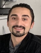 Ein Mann mit dunklem Haar und Bart trägt eine schwarze Jacke und sitzt vor einem unscharfen Hintergrund.