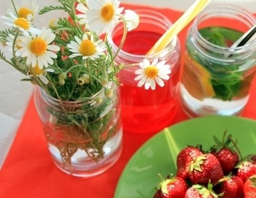 Ein Tisch mit einer roten Tischdecke, auf dem ein Glas mit Kamillenblüten, ein Getränk mit Minze und Strohhalmen sowie eine Schale mit Erdbeeren zu sehen sind.