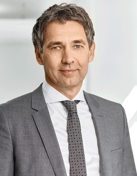 Ein Mann in einem grauen Anzug und Krawatte steht vor einem neutralen Hintergrund.