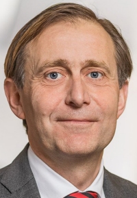 Ein Mann mit kurzen braunen Haaren, blauen Augen und einem ernsten Gesichtsausdruck trägt einen Anzug mit Krawatte.