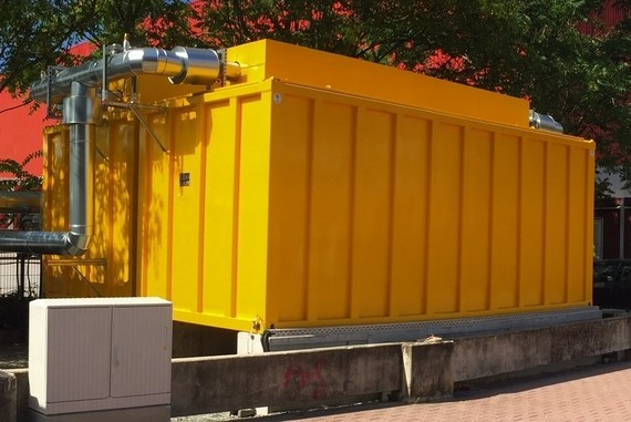 Ein gelber Container steht auf einem Betonsockel im Freien, mit einem roten Hintergrund und Bäumen im Hintergrund.