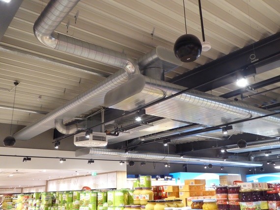 Blick auf die Decke eines Supermarktes mit Rohren, Beleuchtung und Regalen im Hintergrund.