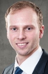 Ein Mann mit blondem Haar trägt einen dunklen Anzug, ein weißes Hemd und eine blaue Krawatte.