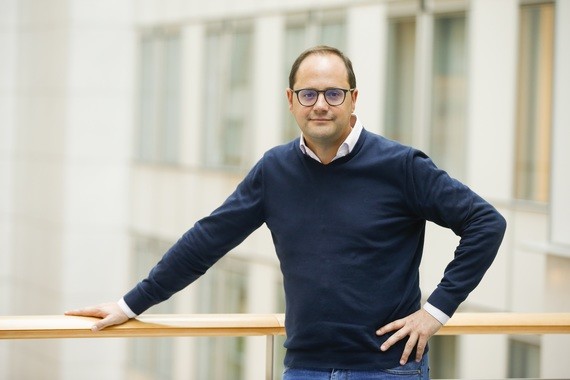 Ein Mann mit Brille und Pullover lehnt lässig an einem Geländer vor einem Gebäude.