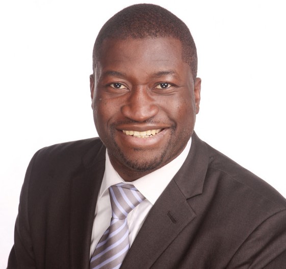 Ein Mann mit dunkler Haut trägt einen dunklen Anzug, ein weißes Hemd und eine blaue Krawatte, sitzt vor einem neutralen Hintergrund.