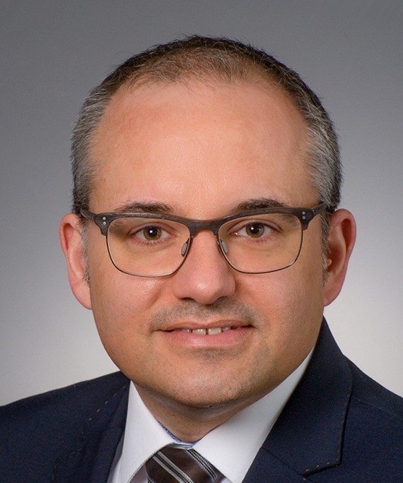 Ein Mann mit Brille, Anzug und Krawatte lächelt in die Kamera.