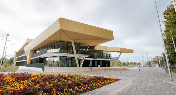 Ein modernes Gebäude mit gelbem Dach und großen Fenstern, umgeben von einem gepflegten Blumenbeet und einem Gehweg.