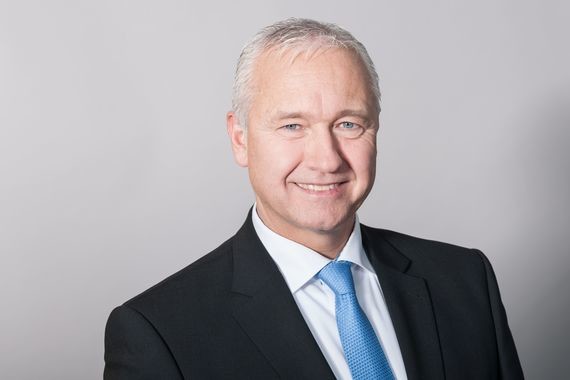 Ein Mann im Anzug und Krawatte lächelt vor einem neutralen Hintergrund.