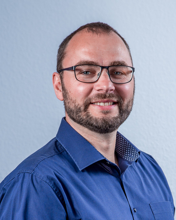 Ein Mann mit Brille und Bart trägt ein blaues Hemd und lächelt in die Kamera.