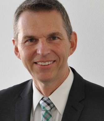 Ein Mann mit kurzen braunen Haaren, einem weißen Hemd, einer grün-weißen Krawatte und einem dunklen Anzug lächelt in die Kamera.