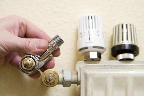Eine Hand hält einen Rohrverschluss, während im Hintergrund ein Heizkörper mit Thermostatkopf zu sehen ist.