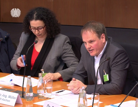 Zwei Personen sitzen an einem Tisch mit Mikrofonen und Dokumenten, während sie an einer offiziellen Veranstaltung teilnehmen.