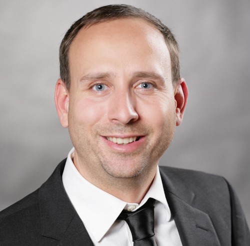 Ein Mann im Anzug und Krawatte lächelt in die Kamera vor einem neutralen Hintergrund.