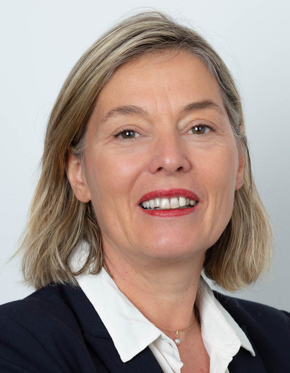 Eine Frau mit kurzen blonden Haaren, lächelnd, trägt ein weißes Hemd und einen dunklen Blazer.