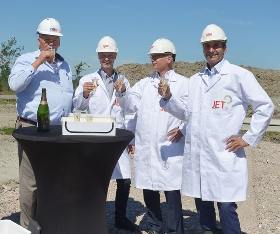 Vier Personen in weißen Laborkitteln und Helmen stehen vor einem Tisch mit Kuchen und Flasche, wobei eine Person das Logo 